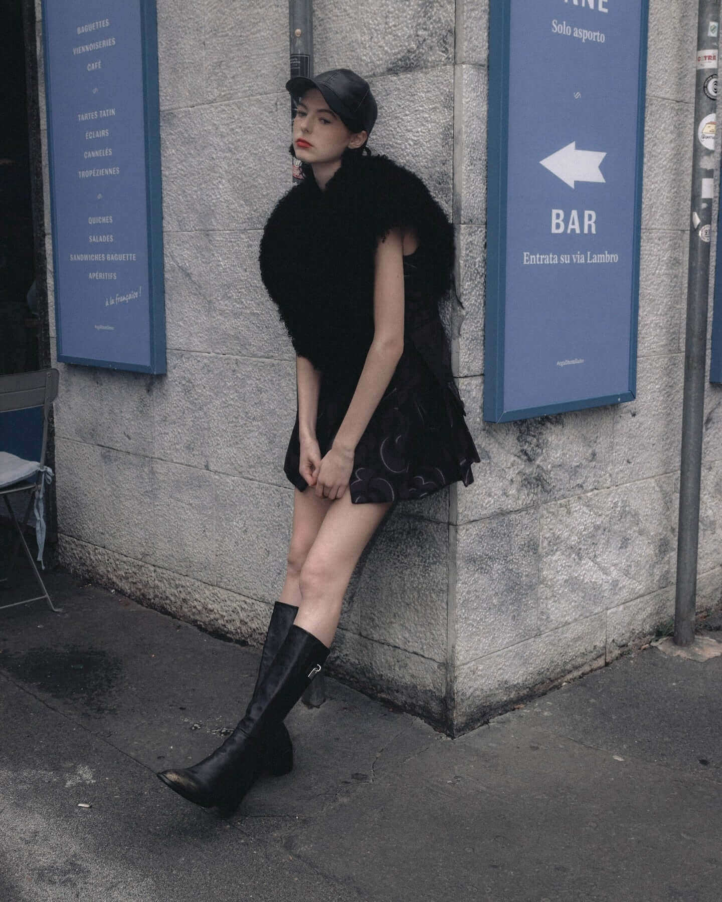 Fashionable young woman in a stylish outfit, wearing tall boots and a fur shawl, leaning against a wall.