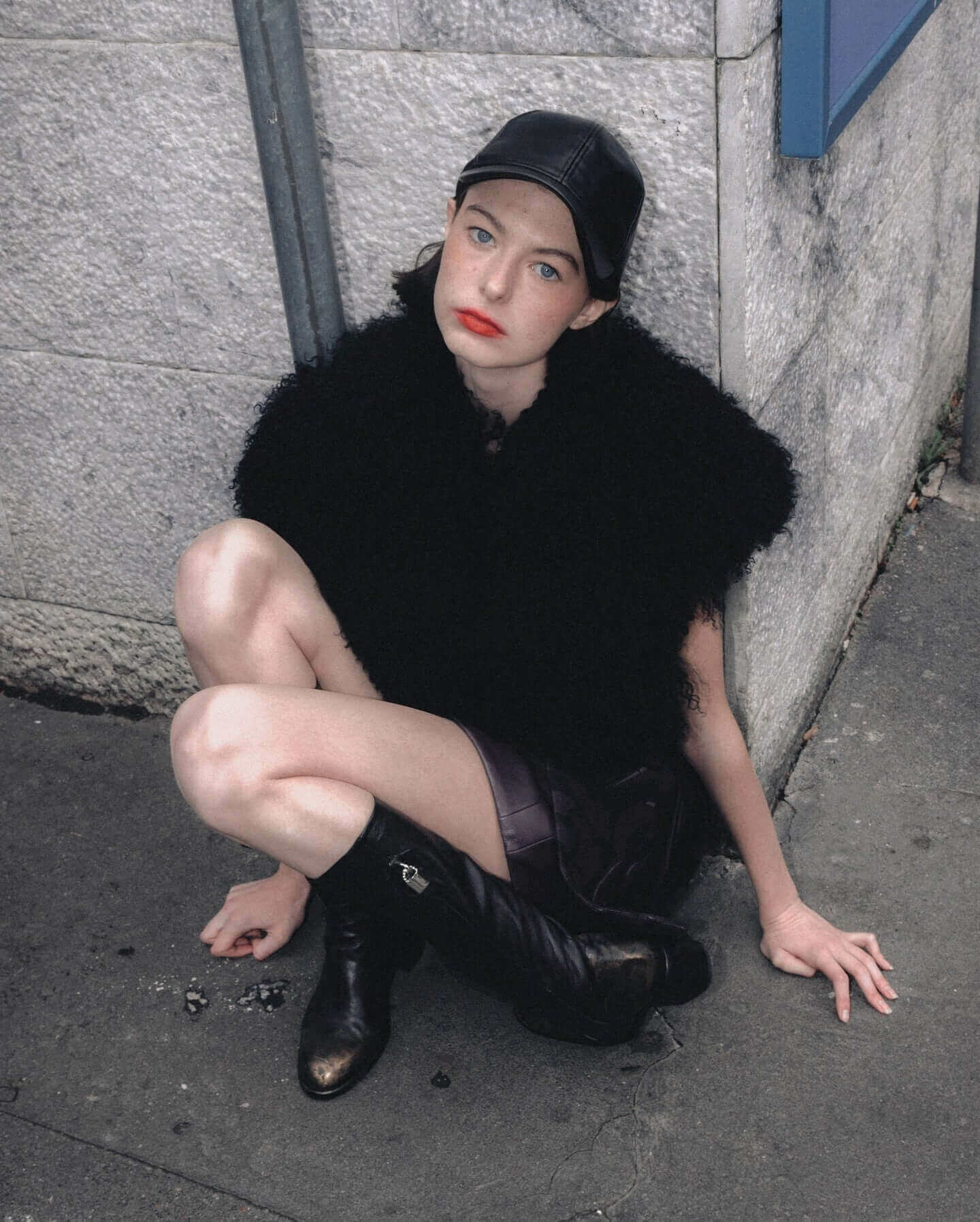 Young model in black fur shawl and leather cap, sitting stylishly against a stone wall on urban street.