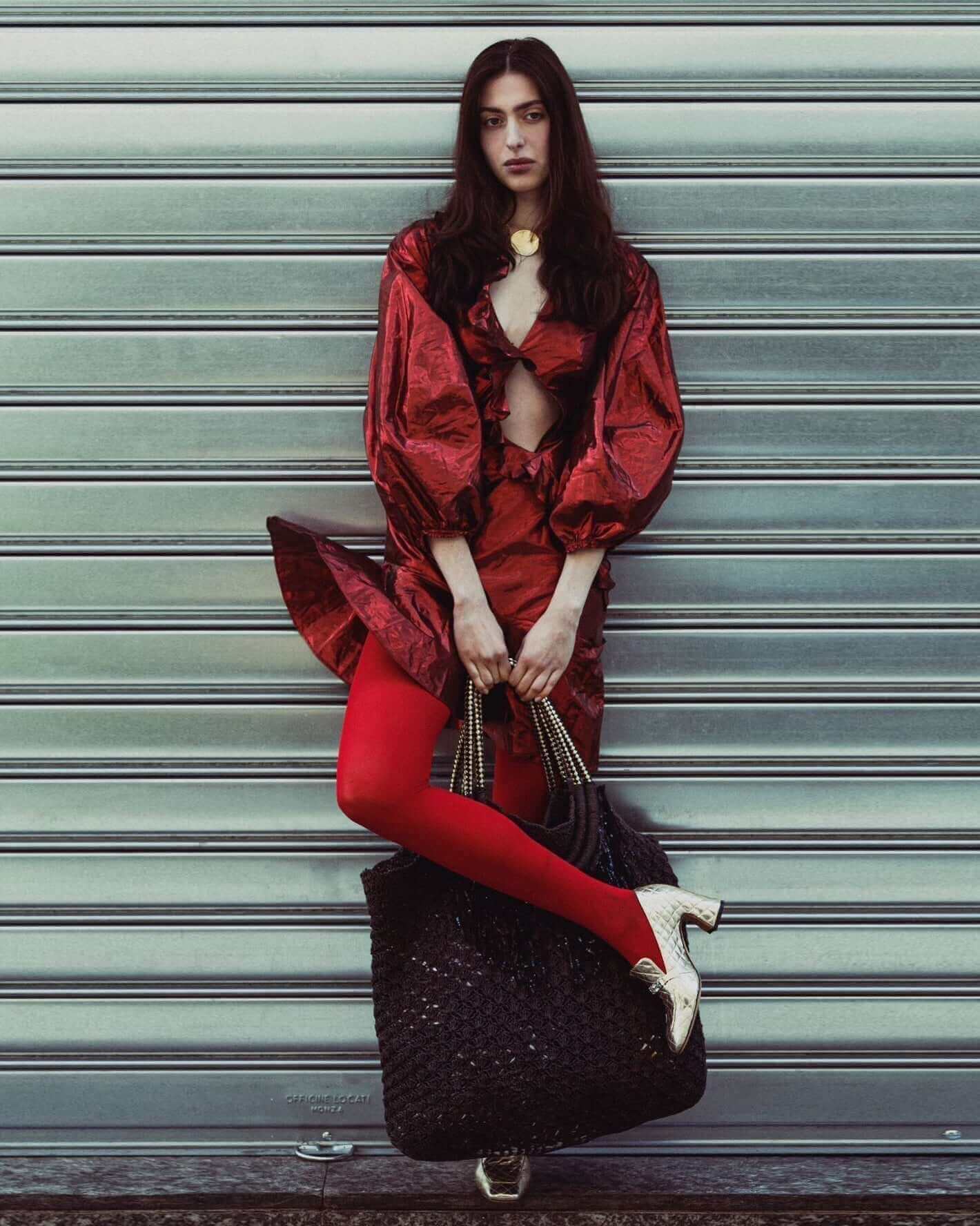 Fashion model in a striking red outfit with bold accessories, posing against a metallic backdrop.