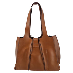A mid-brown polished leather handbag with a wide opening, two top handles, a horizontal gold bar accent on the front, and a matching small brown pouch resting in front of it.