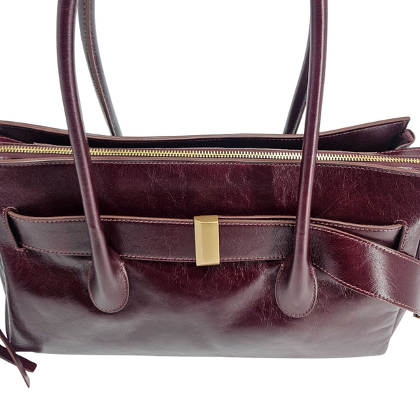 A sleek, burgundy polished leather handbag with two slender top handles and a horizontal gold bar accent on the front.