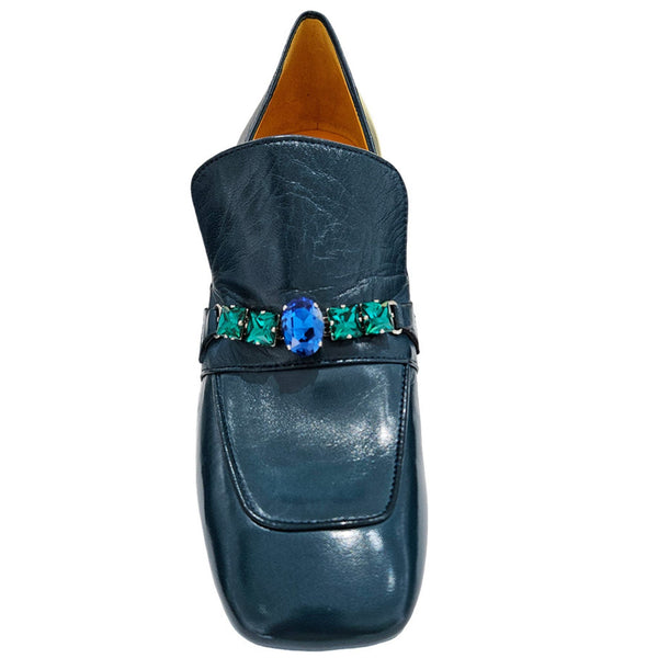 Blue loafers with decorative band featuring green and blue stones on a white background