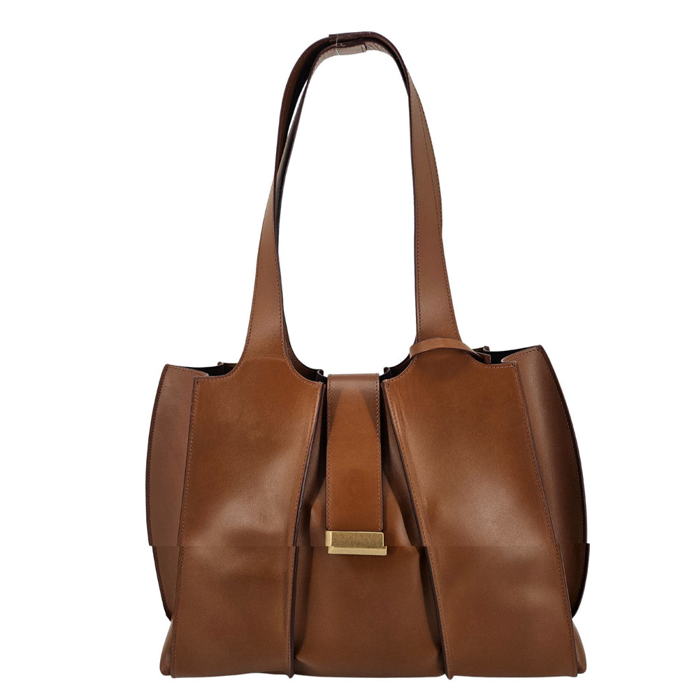 A mid-brown polished leather handbag with a wide opening, two top handles, a horizontal gold bar accent on the front, and a matching small brown pouch resting in front of it.