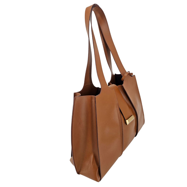 A mid-brown polished leather handbag with a wide opening, two top handles, a horizontal gold bar accent on the front, and a matching small brown pouch resting in front of it.