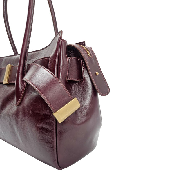 A sleek, burgundy polished leather handbag with two slender top handles and a horizontal gold bar accent on the front.