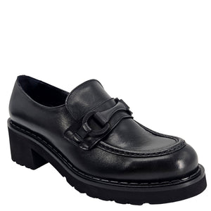 A close-up, eye-level shot of a single black leather lug sole loafer. The shoe features a chunky black lug sole with visible black stitching along the welt. A matching black rubber horse bit buckle adorns the vamp. The leather has a subtle pebbled texture. The background is solid white, emphasizing the shoe.