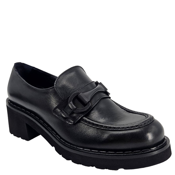 A close-up, eye-level shot of a single black leather lug sole loafer. The shoe features a chunky black lug sole with visible black stitching along the welt. A matching black rubber horse bit buckle adorns the vamp. The leather has a subtle pebbled texture. The background is solid white, emphasizing the shoe.