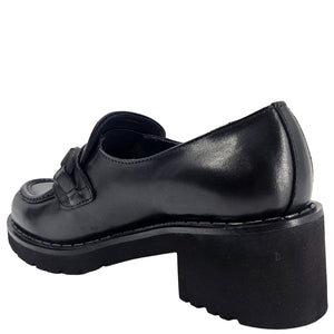A close-up, eye-level shot of a single black leather lug sole loafer. The shoe features a chunky black lug sole with visible black stitching along the welt. A matching black rubber horse bit buckle adorns the vamp. The leather has a subtle pebbled texture. The background is solid white, emphasizing the shoe.