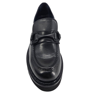 A close-up, eye-level shot of a single black leather lug sole loafer. The shoe features a chunky black lug sole with visible black stitching along the welt. A matching black rubber horse bit buckle adorns the vamp. The leather has a subtle pebbled texture. The background is solid white, emphasizing the shoe.
