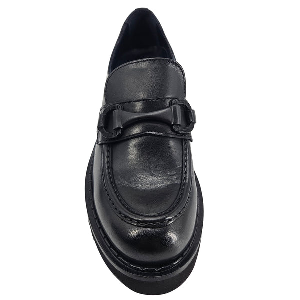 A close-up, eye-level shot of a single black leather lug sole loafer. The shoe features a chunky black lug sole with visible black stitching along the welt. A matching black rubber horse bit buckle adorns the vamp. The leather has a subtle pebbled texture. The background is solid white, emphasizing the shoe.