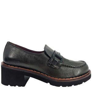 A close-up, eye-level shot of a single dark green leather lug sole loafer. The shoe features a chunky black lug sole with a visible brown welt stitching. A dark green rubber horse bit buckle adorns the vamp. The leather has a subtle pebbled texture. The background is solid white, emphasizing the shoe.