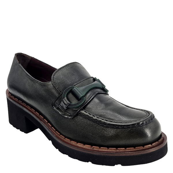 A close-up, eye-level shot of a single dark green leather lug sole loafer. The shoe features a chunky black lug sole with a visible brown welt stitching. A dark green rubber horse bit buckle adorns the vamp. The leather has a subtle pebbled texture. The background is solid white, emphasizing the shoe.