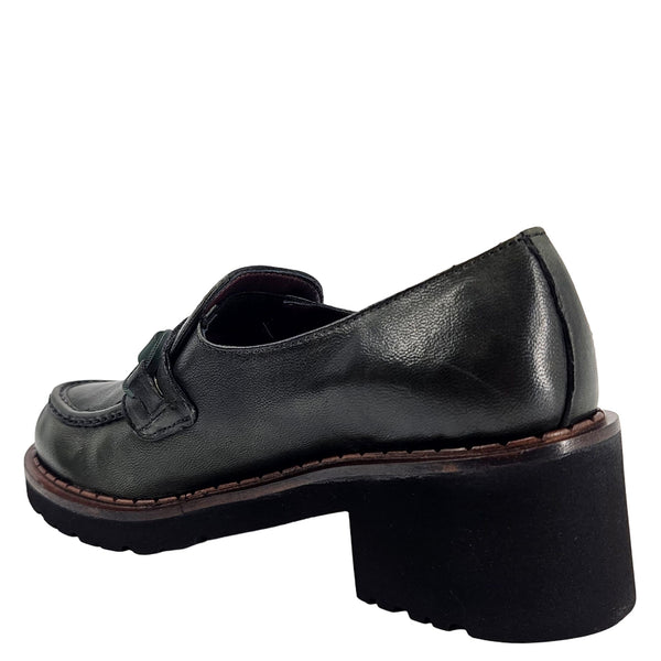 A close-up, eye-level shot of a single dark green leather lug sole loafer. The shoe features a chunky black lug sole with a visible brown welt stitching. A dark green rubber horse bit buckle adorns the vamp. The leather has a subtle pebbled texture. The background is solid white, emphasizing the shoe.