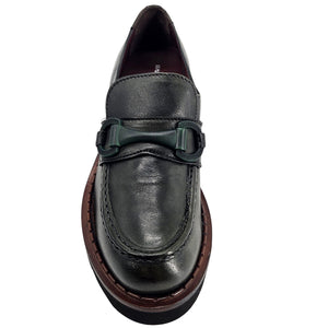 A close-up, eye-level shot of a single dark green leather lug sole loafer. The shoe features a chunky black lug sole with a visible brown welt stitching. A dark green rubber horse bit buckle adorns the vamp. The leather has a subtle pebbled texture. The background is solid white, emphasizing the shoe.