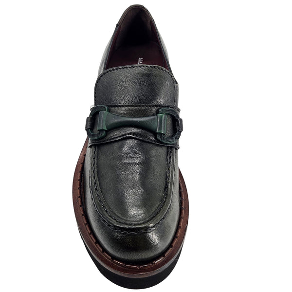 A close-up, eye-level shot of a single dark green leather lug sole loafer. The shoe features a chunky black lug sole with a visible brown welt stitching. A dark green rubber horse bit buckle adorns the vamp. The leather has a subtle pebbled texture. The background is solid white, emphasizing the shoe.