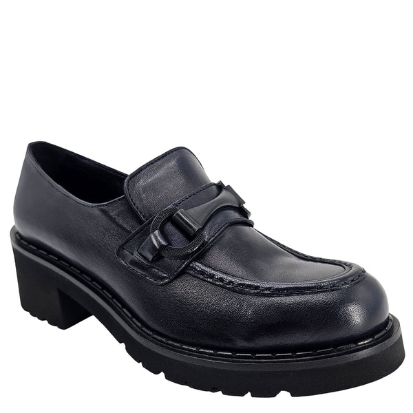 A close-up, eye-level shot of a single dark navy blue leather lug sole loafer. The shoe features a chunky black lug sole with visible dark stitching along the welt. A matching dark navy blue rubber horse bit buckle adorns the vamp. The leather has a subtle pebbled texture. The background is solid white, emphasizing the shoe.