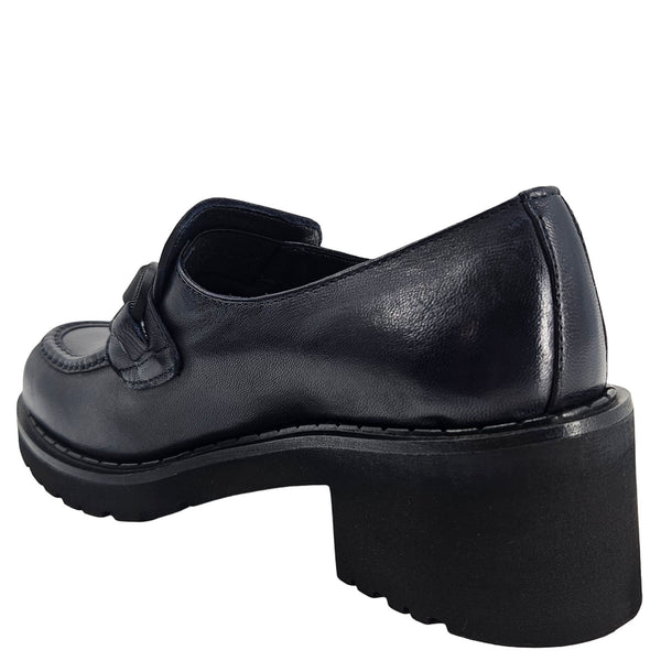 A close-up, eye-level shot of a single dark navy blue leather lug sole loafer. The shoe features a chunky black lug sole with visible dark stitching along the welt. A matching dark navy blue rubber horse bit buckle adorns the vamp. The leather has a subtle pebbled texture. The background is solid white, emphasizing the shoe.