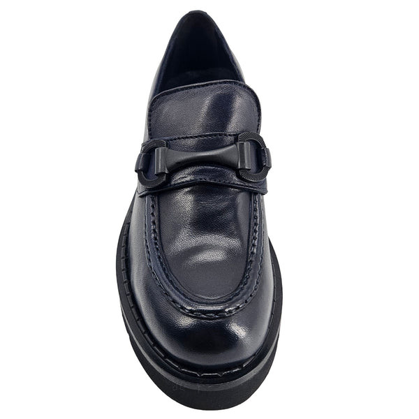 A close-up, eye-level shot of a single dark navy blue leather lug sole loafer. The shoe features a chunky black lug sole with visible dark stitching along the welt. A matching dark navy blue rubber horse bit buckle adorns the vamp. The leather has a subtle pebbled texture. The background is solid white, emphasizing the shoe.
