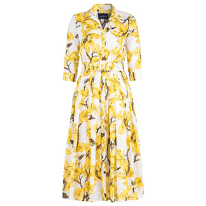 A white midi dress with a vibrant yellow magnolia blossom print. The dress features a shirt collar, 3/4 sleeves, a button-front, and a pleated A-line skirt with a matching fabric belt.