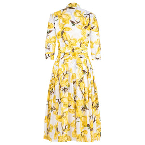 A white midi dress with a vibrant yellow magnolia blossom print. The dress features a shirt collar, 3/4 sleeves, a button-front, and a pleated A-line skirt with a matching fabric belt.