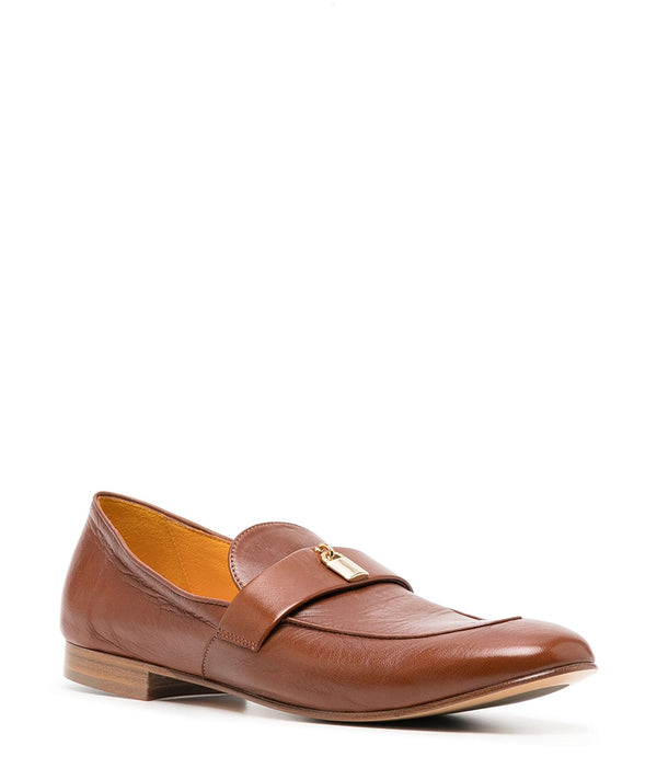 A leather loafer with a flat sole and rounded toe, featuring a small metal lock accent centered on the upper strap. Designed by Madison in Beverly Hills and handcrafted in Italy.