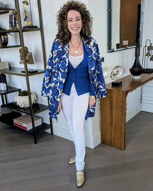 Woman in a blue floral jacket and white pants standing in a room with shelves and decor.