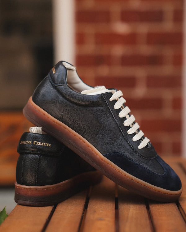Navy blue leather sneaker with white laces on a wooden surface against a brick wall background.