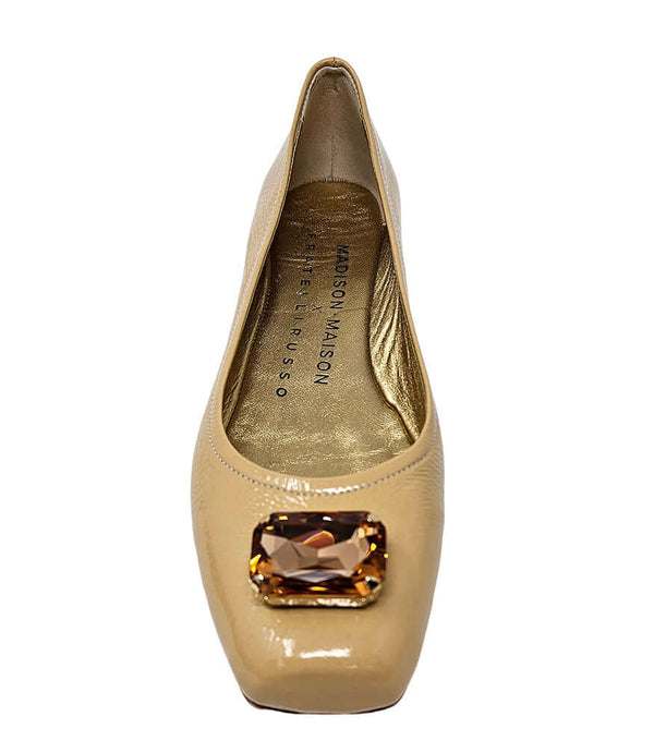 Four-angle view of a women's flat shoe with a glossy leather finish, rounded square toe, and a large faceted jewel embellishment at the toe. The shoe features a low-profile sole and a metallic-toned interior lining, displayed from side, angled, back, and