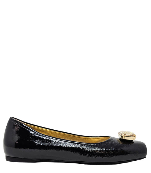 Four-angle view of a women's flat shoe with a glossy leather finish, rounded square toe, and a large faceted jewel embellishment at the toe. The shoe features a low-profile sole and a metallic-toned interior lining, displayed from side, angled, back, and