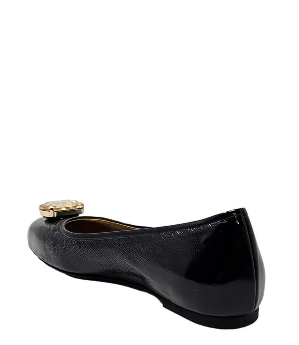Four-angle view of a women's flat shoe with a glossy leather finish, rounded square toe, and a large faceted jewel embellishment at the toe. The shoe features a low-profile sole and a metallic-toned interior lining, displayed from side, angled, back, and