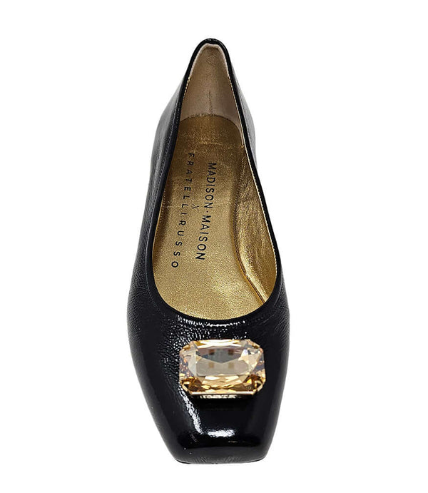 Four-angle view of a women's flat shoe with a glossy leather finish, rounded square toe, and a large faceted jewel embellishment at the toe. The shoe features a low-profile sole and a metallic-toned interior lining, displayed from side, angled, back, and