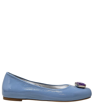 Four-angle view of a women's flat shoe with a glossy leather finish, rounded square toe, and a large faceted jewel embellishment at the toe. The shoe features a low-profile sole and a metallic-toned interior lining, displayed from side, angled, back, and