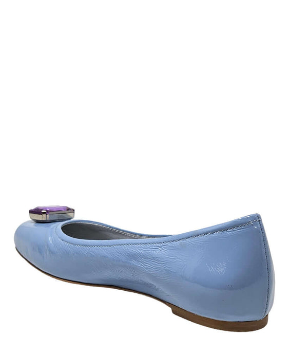 Four-angle view of a women's flat shoe with a glossy leather finish, rounded square toe, and a large faceted jewel embellishment at the toe. The shoe features a low-profile sole and a metallic-toned interior lining, displayed from side, angled, back, and