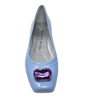 Four-angle view of a women's flat shoe with a glossy leather finish, rounded square toe, and a large faceted jewel embellishment at the toe. The shoe features a low-profile sole and a metallic-toned interior lining, displayed from side, angled, back, and