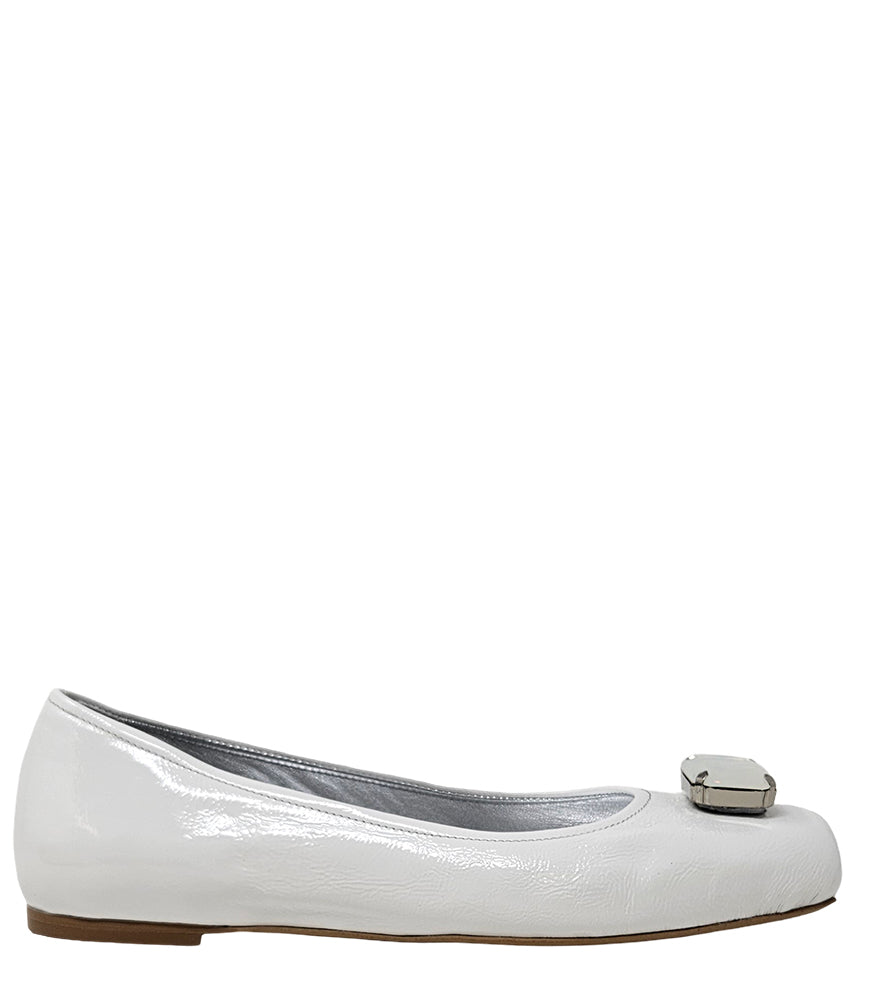 Four-angle view of a women's flat shoe with a glossy leather finish, rounded square toe, and a large faceted jewel embellishment at the toe. The shoe features a low-profile sole and a metallic-toned interior lining, displayed from side, angled, back, and