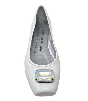 Four-angle view of a women's flat shoe with a glossy leather finish, rounded square toe, and a large faceted jewel embellishment at the toe. The shoe features a low-profile sole and a metallic-toned interior lining, displayed from side, angled, back, and