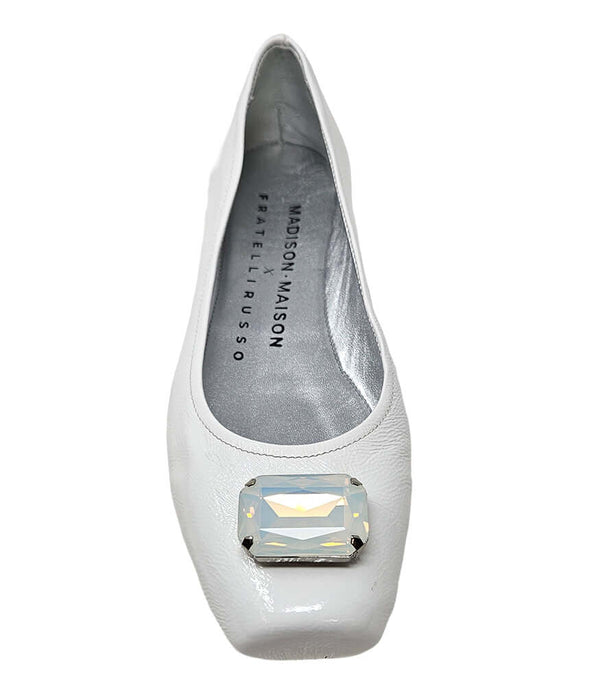 Four-angle view of a women's flat shoe with a glossy leather finish, rounded square toe, and a large faceted jewel embellishment at the toe. The shoe features a low-profile sole and a metallic-toned interior lining, displayed from side, angled, back, and