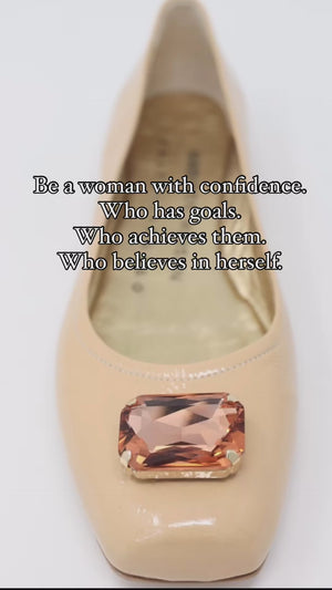 Four-angle view of a women's flat shoe with a glossy leather finish, rounded square toe, and a large faceted jewel embellishment at the toe. The shoe features a low-profile sole and a metallic-toned interior lining, displayed from side, angled, back, and