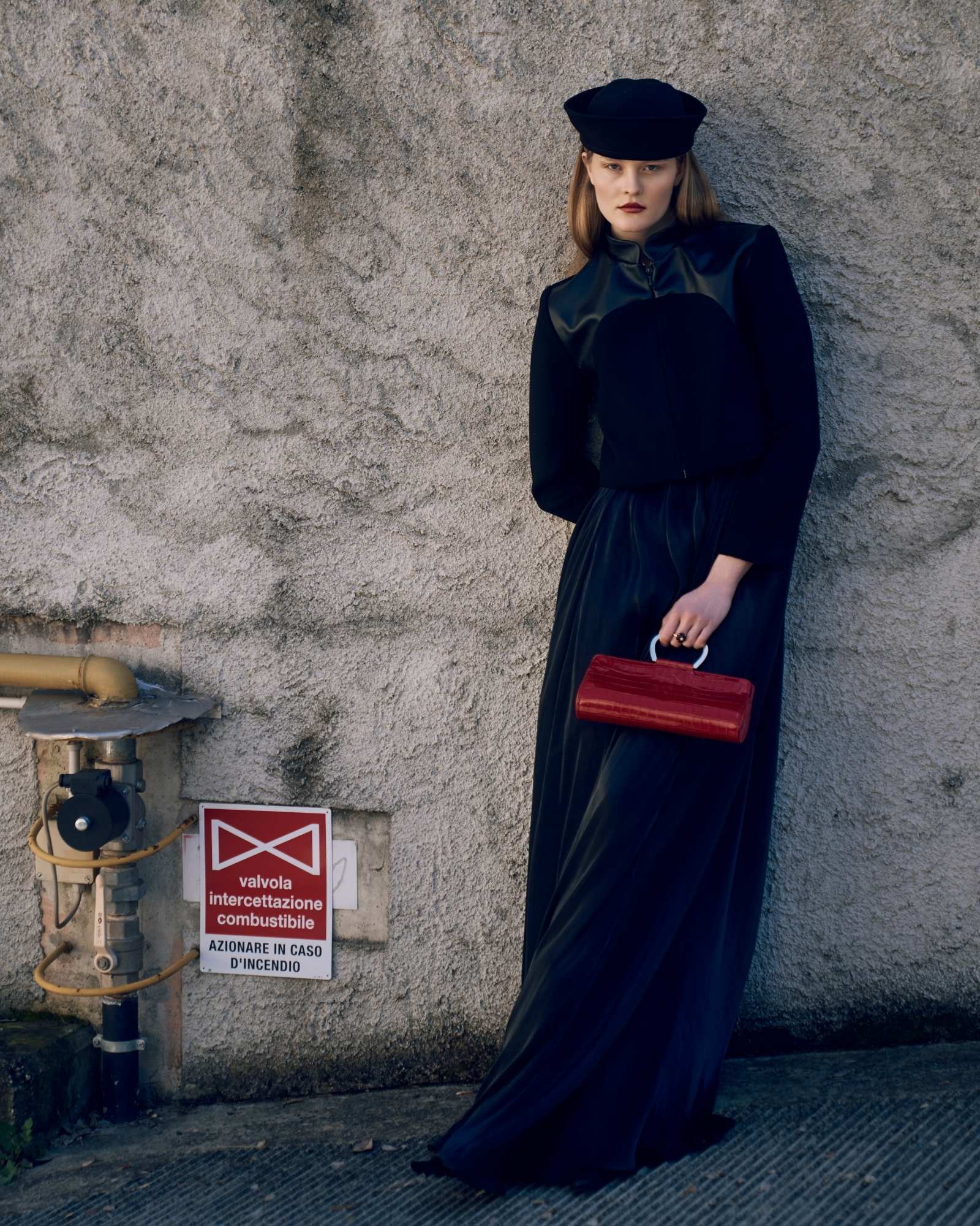 Woman in a long dark coat and red handbag against a textured wall.