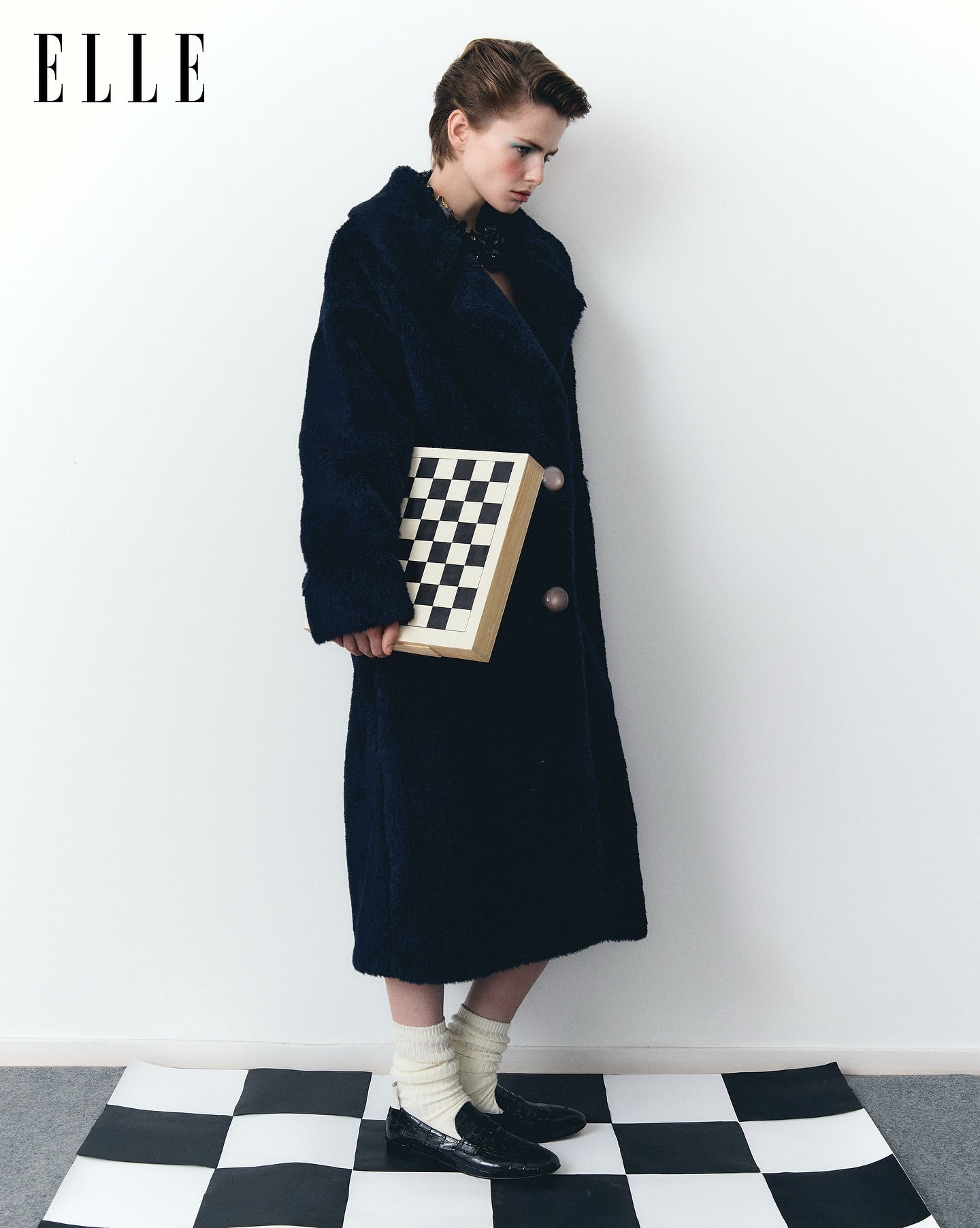 Person wearing a long dark coat holding a checkered bag on a black and white checkered floor with 'ELLE' branding.