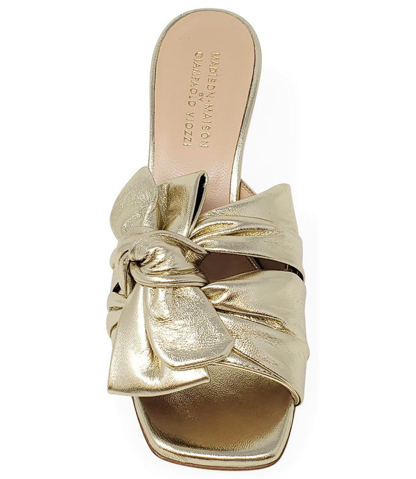 Open-toe mule handcrafted in Italy, featuring a metallic finish, wide vamp with a twisted bow detail, and a flared heel. Shown from multiple angles against a clean background