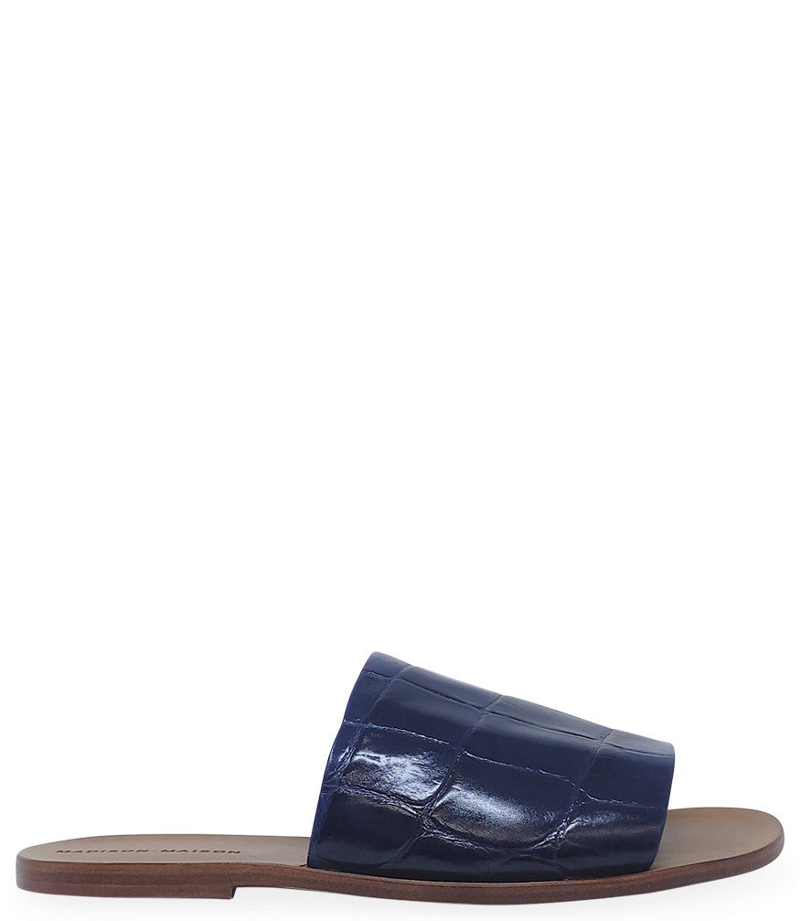 Handmade slide sandal crafted in Italy with a wide, textured band across the foot and a flat leather sole. Features a structured, embossed finish. Displayed from front and side angles against a clean background.