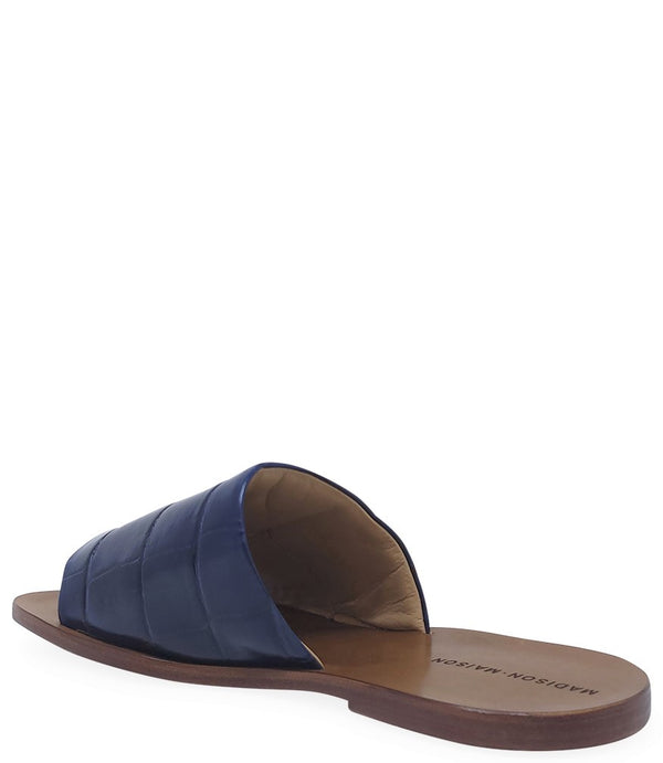 Handmade slide sandal crafted in Italy with a wide, textured band across the foot and a flat leather sole. Features a structured, embossed finish. Displayed from front and side angles against a clean background.